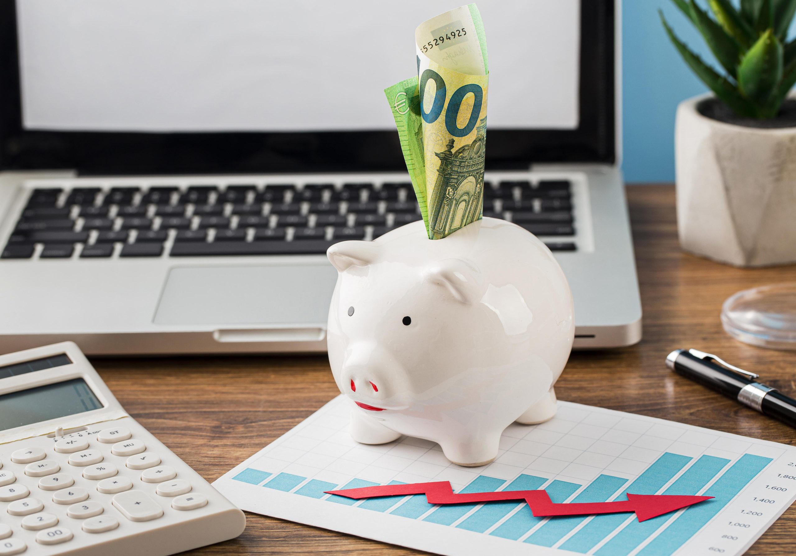 A white piggy bank with euro bills sticking out sits on a financial chart with upward blue bars and a red arrow, placed in front of an open laptop. A calculator and pen rest nearby, symbolizing savings growth, investment planning, and 401(k) retirement contributions in a modern workspace.