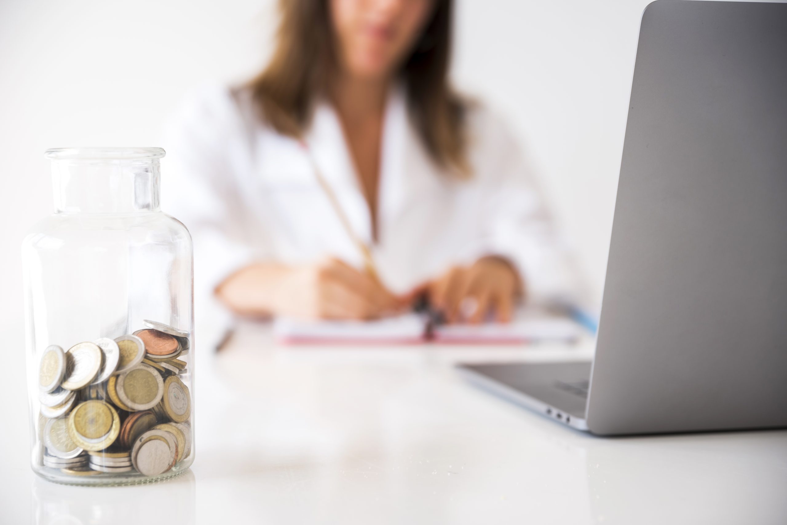 A close-up of a glass jar filled with assorted coins on a white desk, with a blurred woman in a lab coat working on paperwork beside a laptop in the background, symbolizing the importance of financial precautions in professional and personal planning.