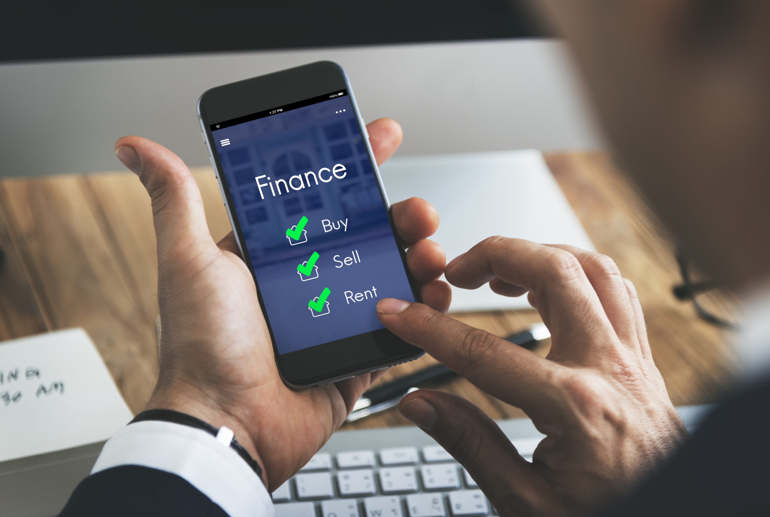 A man in a suit uses a smartphone displaying a finance app with checkmarks next to options for Buy, Sell, and Rent. The setting includes a modern workspace with a keyboard and notepad, symbolizing the use of digital tools to control spending and manage financial decisions efficiently.