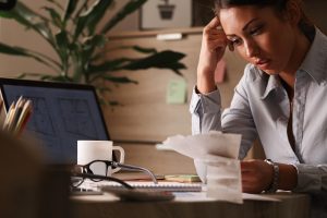 A concerned woman sits at her desk reviewing a stack of receipts and financial documents, with a laptop and coffee mug nearby, illustrating the stress and pressure of managing dollar debts.
