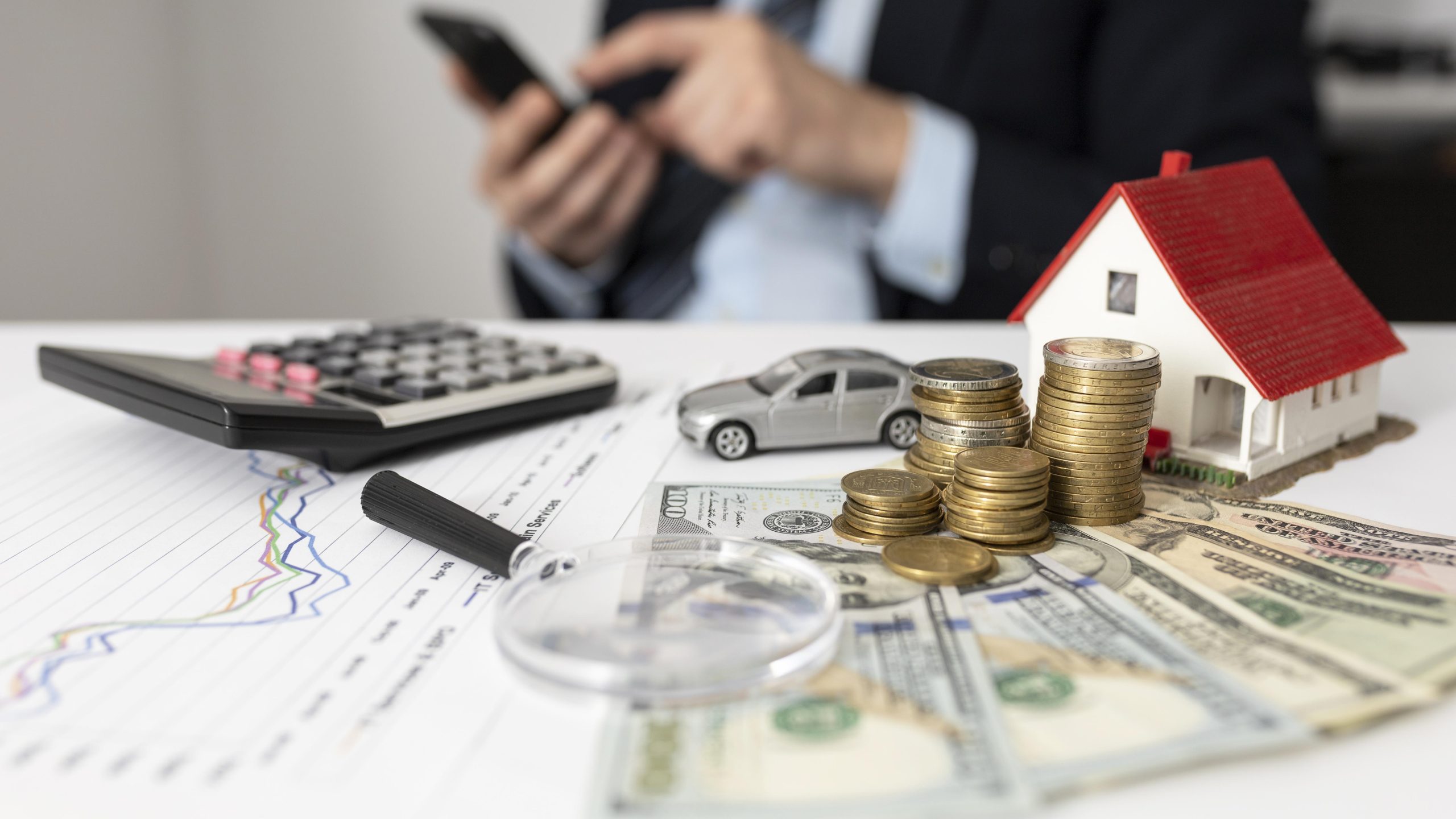 A financial setup on a desk features stacked coins, US dollar bills, a calculator, a toy car, and a miniature house, symbolizing budgeting and asset management in preparation for financial emergencies; a businessperson uses a smartphone in the background.