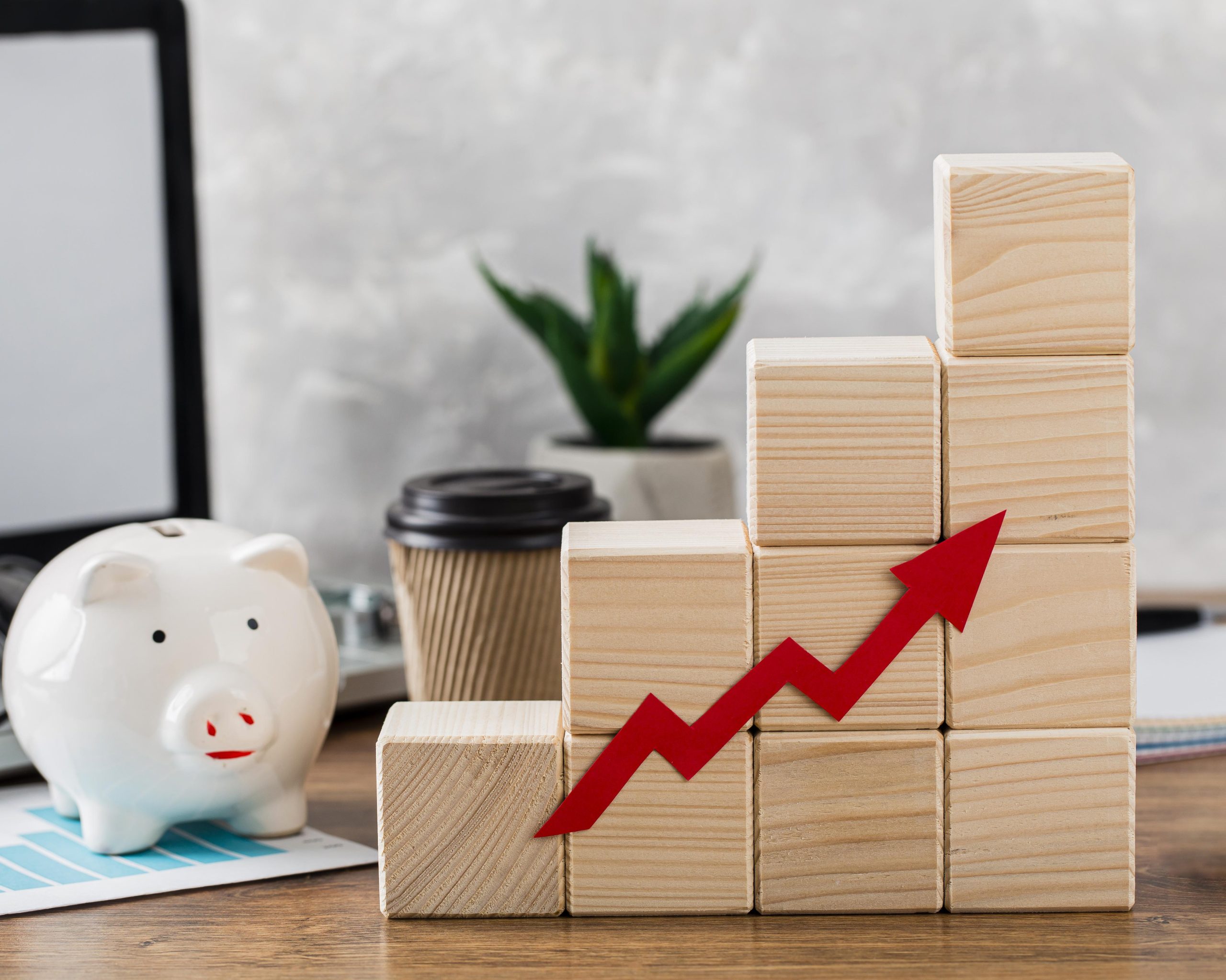 A desk scene shows a white piggy bank beside stacked wooden blocks forming a rising bar graph with a red arrow, symbolizing growth and strategic financial priorities; a laptop, coffee cup, and plant appear in the background.