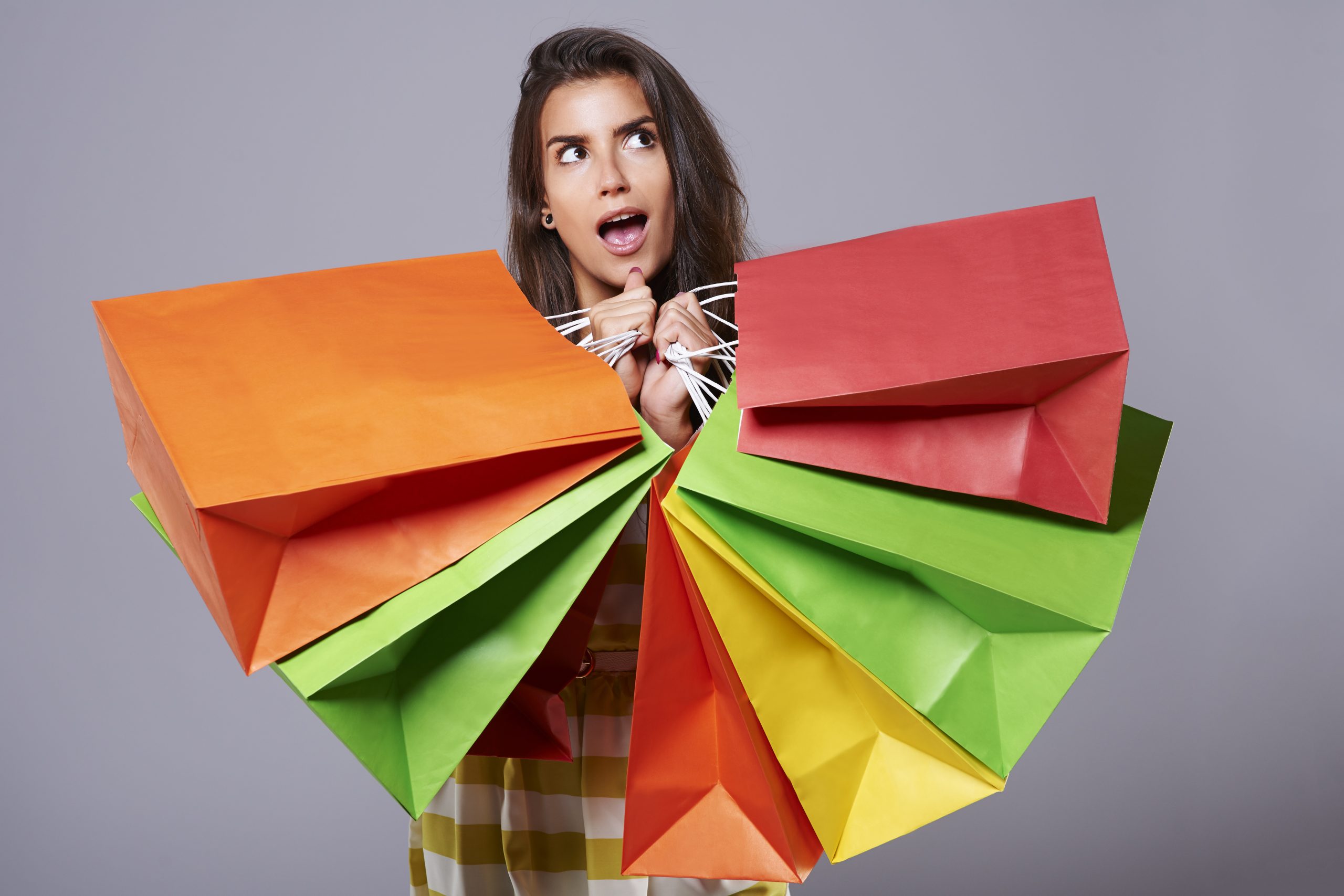 A surprised young woman holds a large number of colorful shopping bags in both hands, suggesting a recent shopping spree and visually representing the concept of impulsive purchases driven by emotion or sudden desire.
