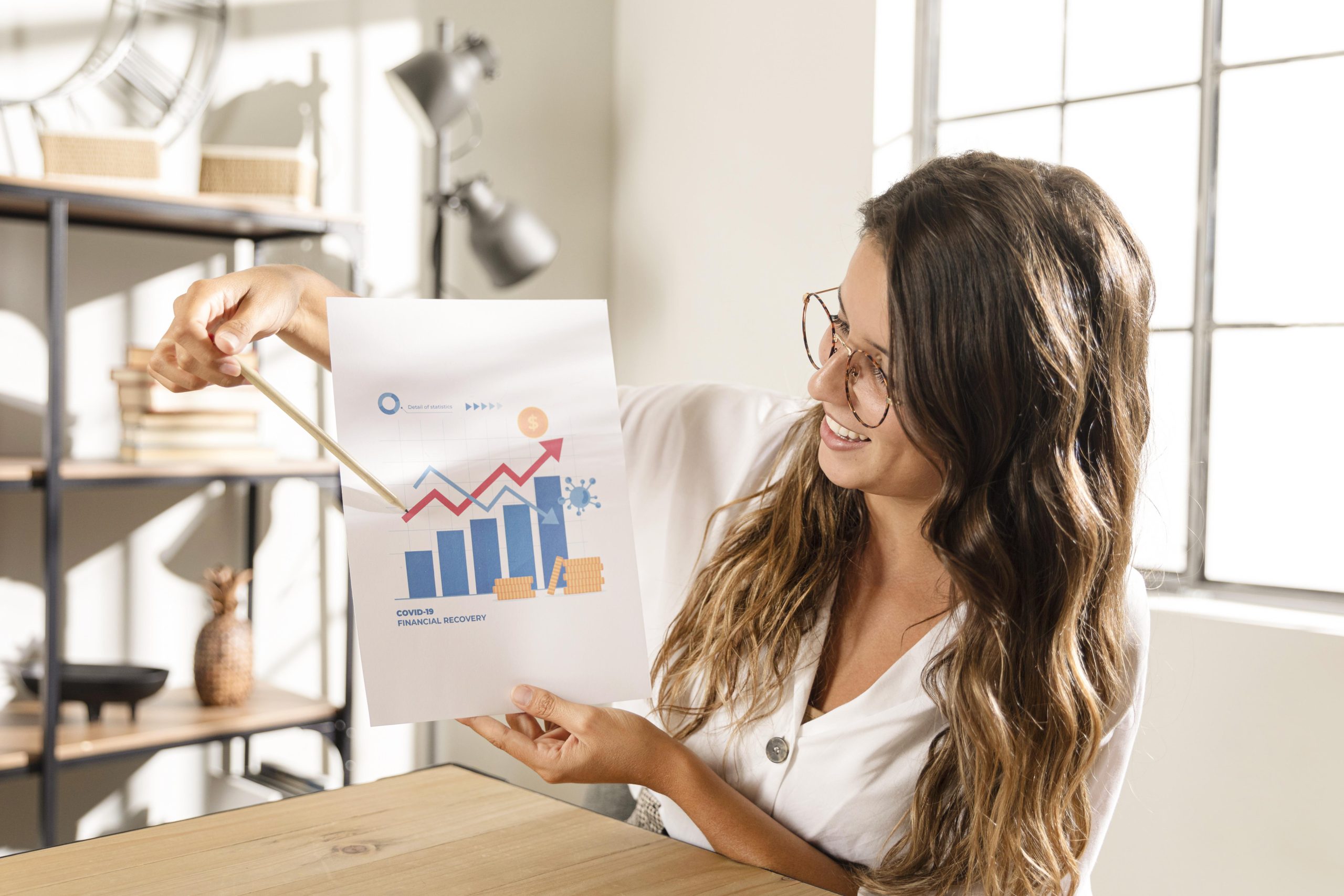 A smiling woman in glasses and a white blouse holds up a chart labeled "COVID-19 Financial Recovery" and points at it with a pencil. The colorful graph includes upward-trending bars and lines, symbolizing measurable financial progress and recovery in a bright, modern office setting.