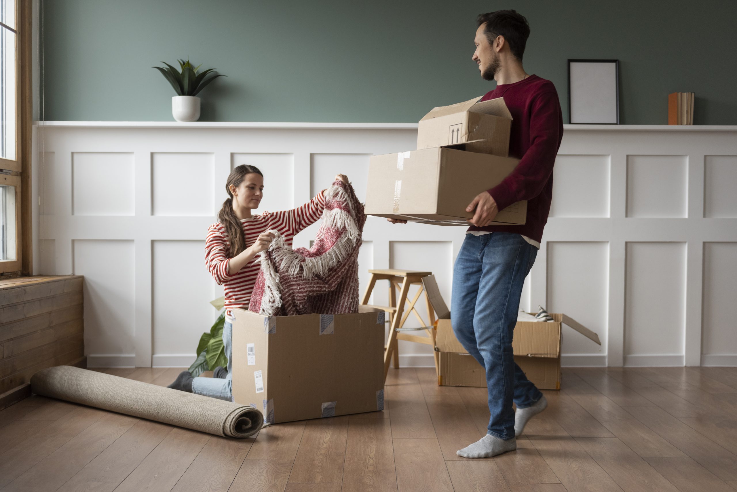 A couple is unpacking boxes in their new home, with the woman kneeling to organize a blanket and the man carrying cardboard boxes, illustrating the importance of planning ahead to prepare financially for a major life transition like moving.