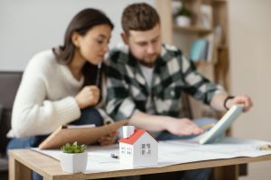 A couple sits together at a table reviewing documents and architectural plans, with a small model house in the foreground, representing the process of refinancing a property and making informed financial decisions.