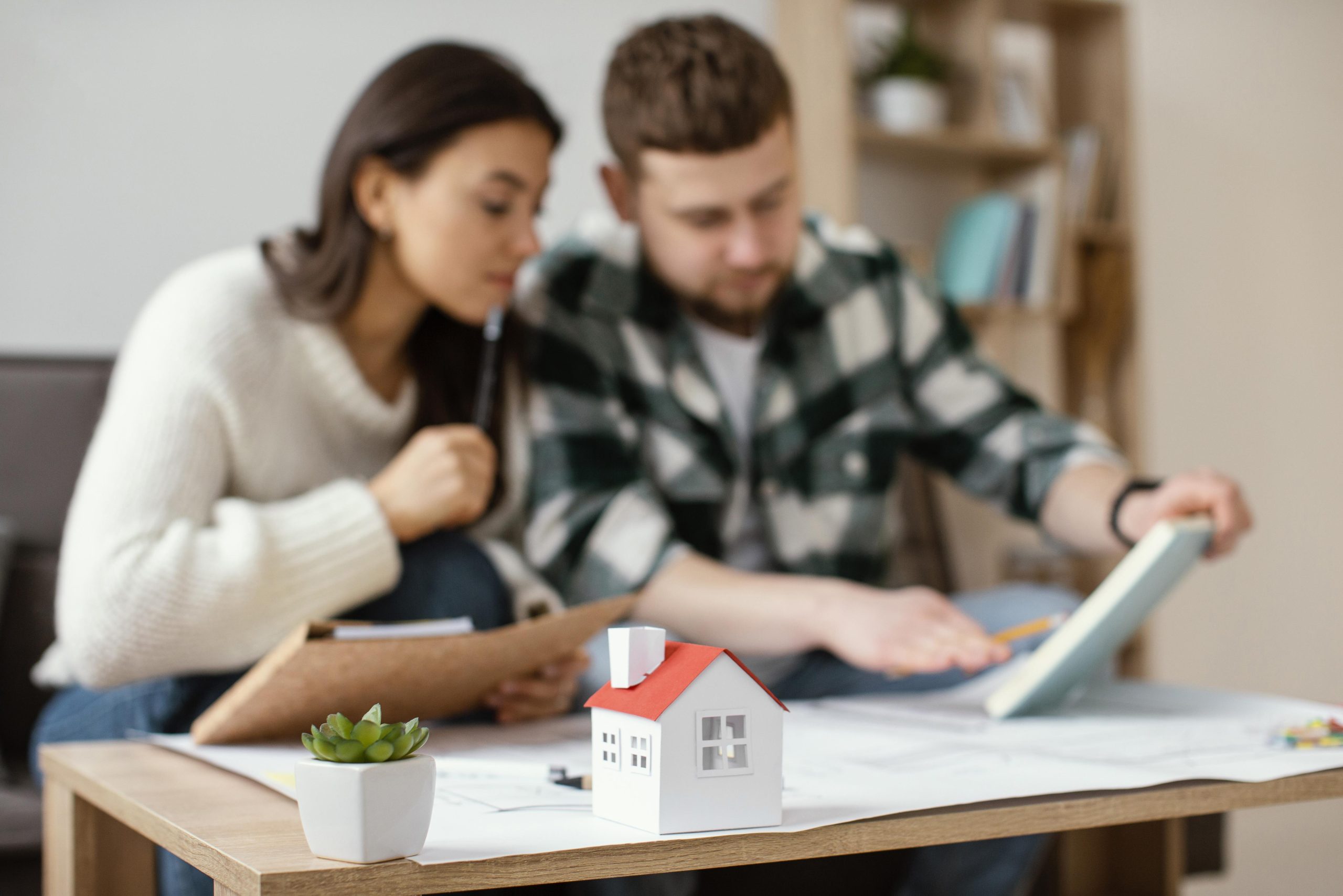 A couple sits together at a table reviewing documents and architectural plans, with a small model house in the foreground, representing the process of refinancing a property and making informed financial decisions.