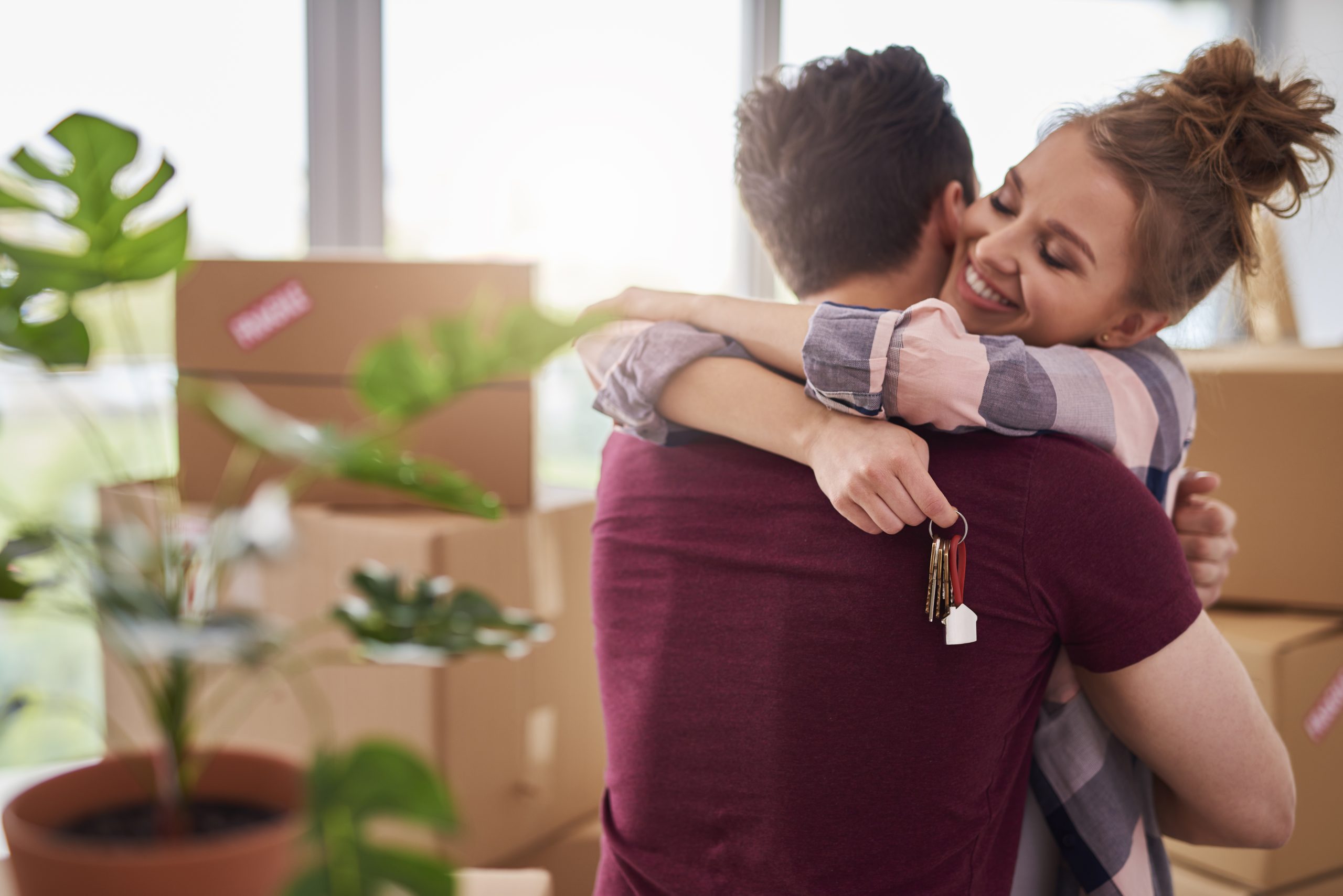 A joyful young couple embraces in a sunlit apartment filled with moving boxes and green plants, celebrating a fresh start after renting or buying a property; the woman smiles while holding a keychain with a small house charm in her hand.