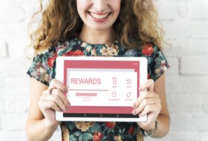 A smiling woman in a floral dress holds a tablet displaying a digital interface with the word "REWARDS" and icons for dining, travel, recommendations, and lifestyle, representing the benefits of rewards programs in a visually appealing and modern context.