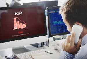 A man talks on the phone at his desk, analyzing financial charts on two computer monitors; one screen displays a downward-trending bar graph labeled "Risk," emphasizing the warning signs that the investment is a scam.