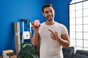 A smiling man in a white t-shirt stands in a brightly lit room, proudly holding up a pink piggy bank and pointing at it, promoting the idea to save money effectively.