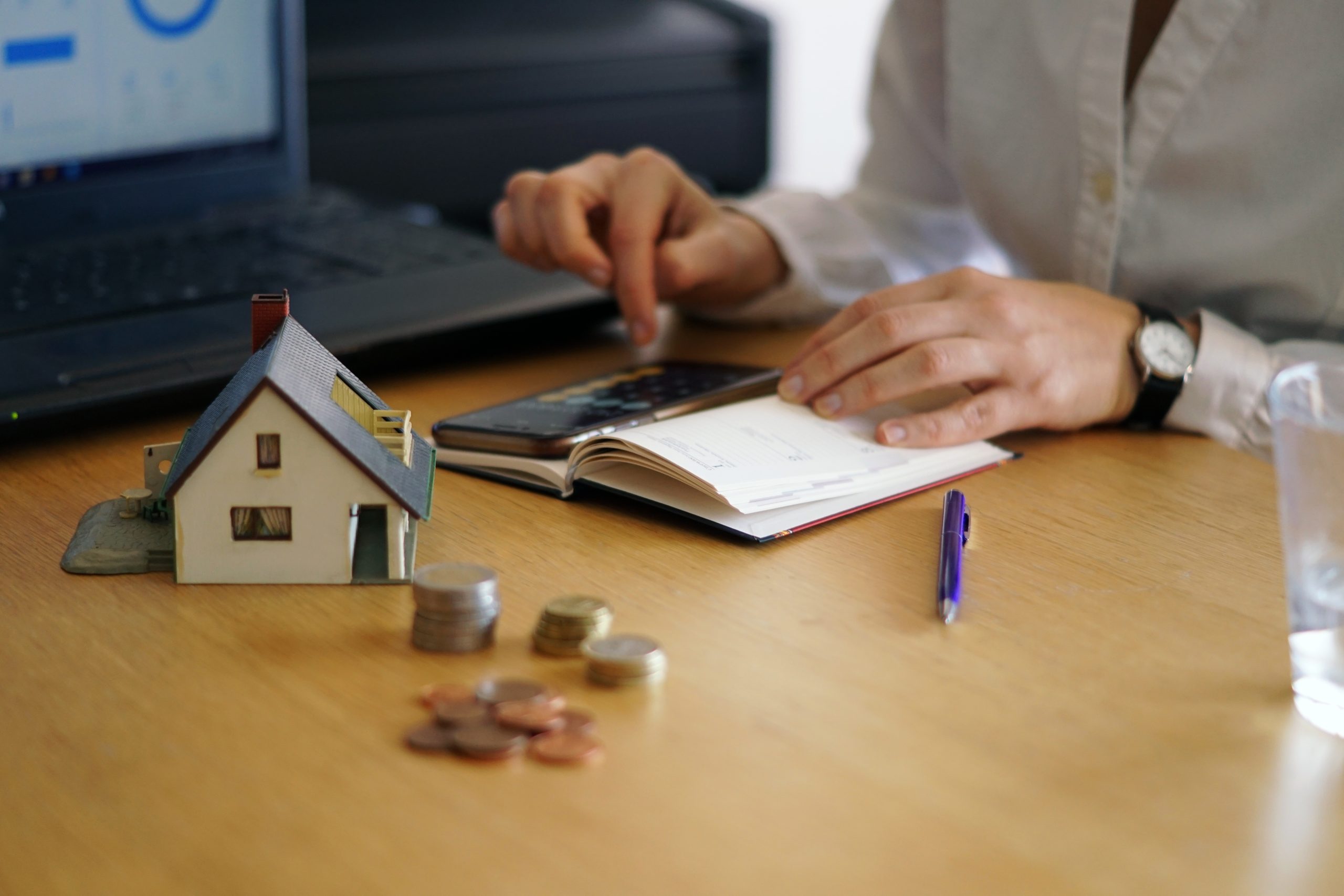 A person sits at a desk reviewing notes with a calculator and planner, surrounded by coins, a miniature house model, and a laptop displaying charts, symbolizing careful planning to determine the best type of financing for a real estate investment or personal budget.
