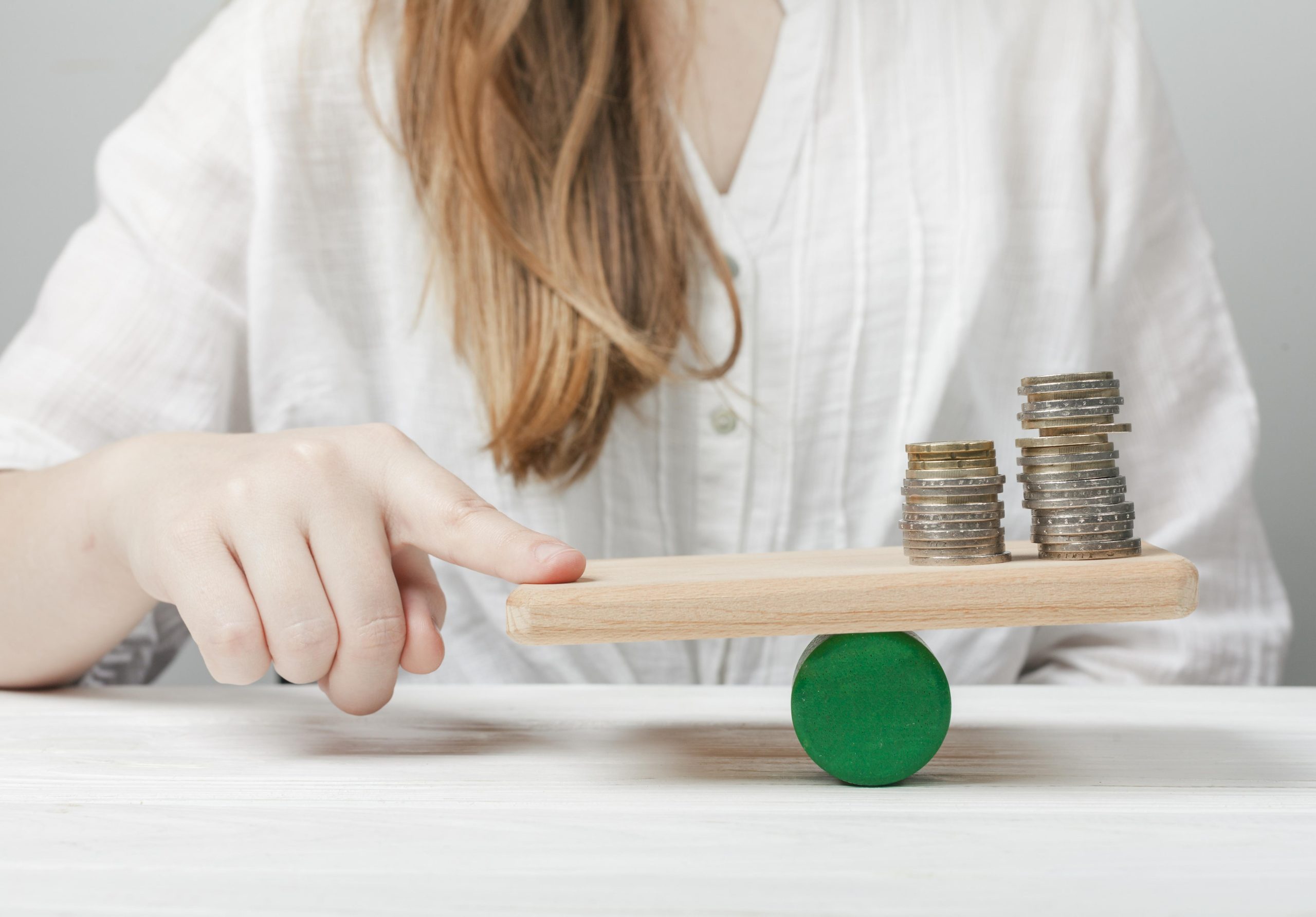 A woman balances a wooden board on a green cylinder with stacks of coins on one end, symbolizing the careful management required to deal with variable income and maintain financial stability.